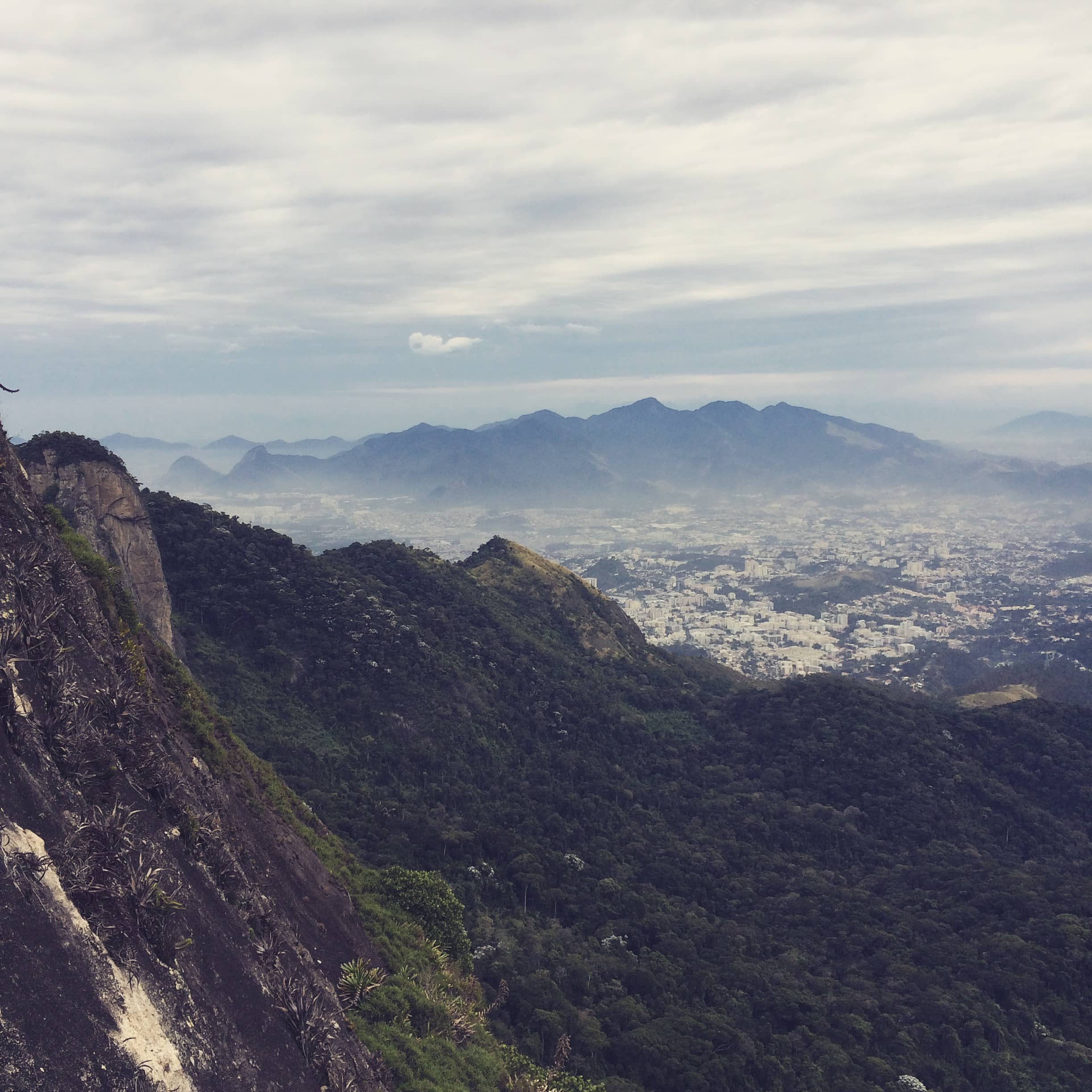 Forested mountain ridges frame a sweeping view of the city beneath an overcast sky.