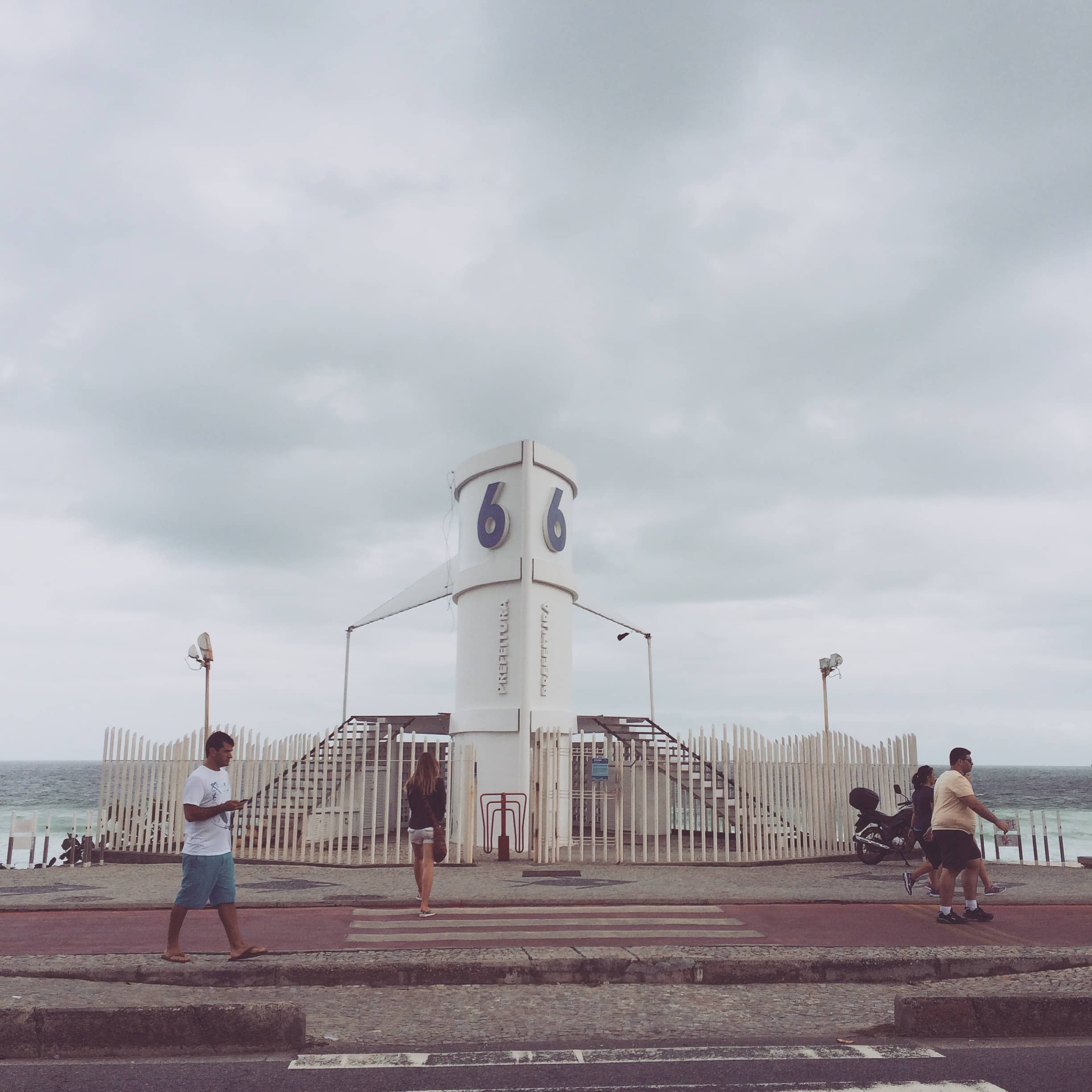 People gather around a white clock tower on a waterfront promenade.