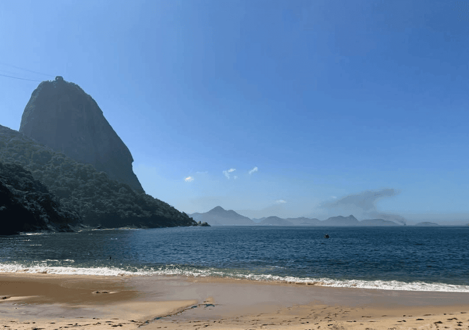 A sandy beach meets blue water beneath a steep, tree-covered mountain.