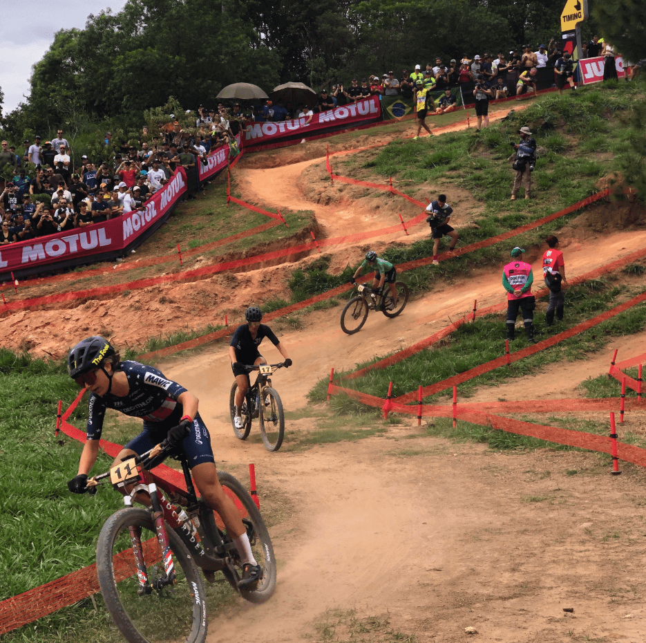 Cyclists lean into a dirt turn as spectators watch from the edge of the course.