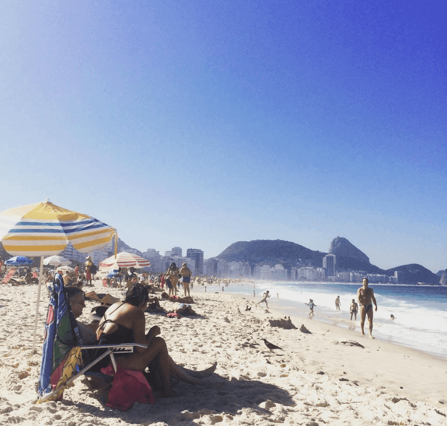 Beachgoers and a striped umbrella line a bright sandy shore with mountains beyond.
