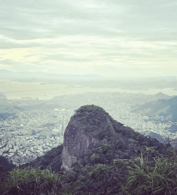 A rounded rock formation rises above forest with a hazy city in the distance.