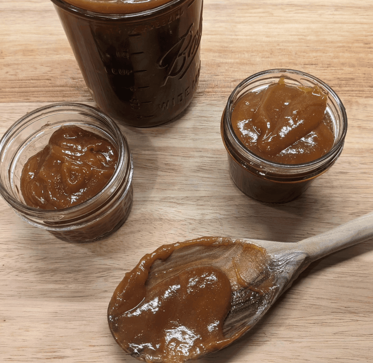 Small jars of amber-colored preserves and a wooden spoon sit on a wooden surface.