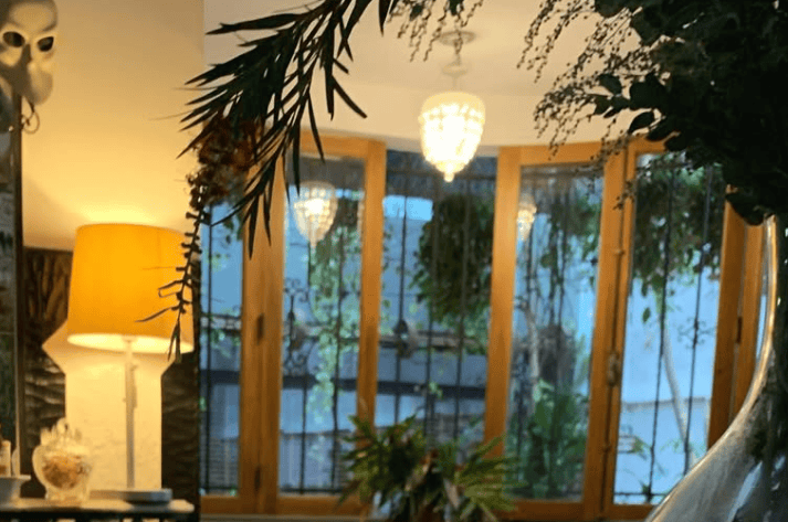 A glowing table lamp and hanging light illuminate a room filled with plants beside glass doors.