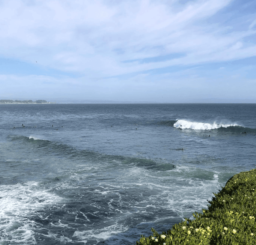 White waves roll toward a rocky coastal edge beneath a pale blue sky.