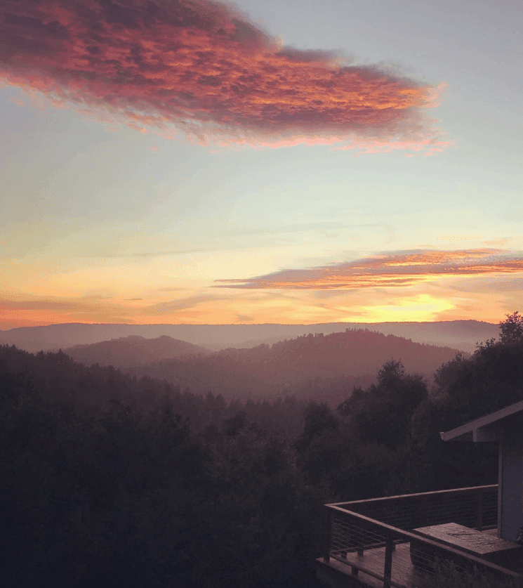 Pink clouds glow above layered hills and a dark tree canopy at sunset.