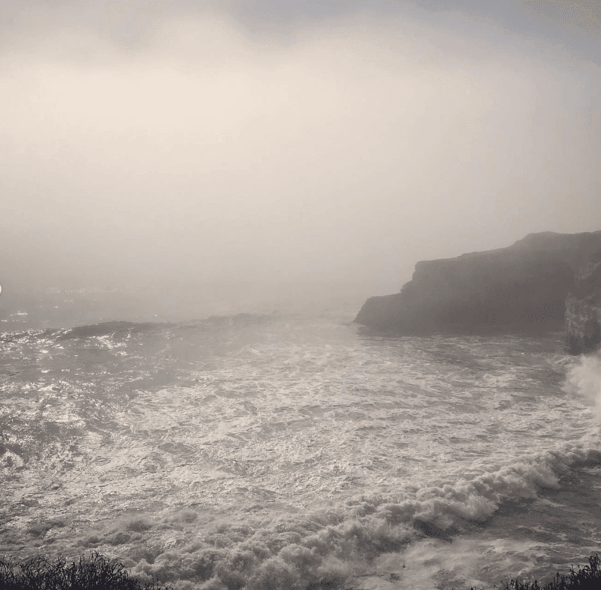 Sunlight reflects off waves beside a rocky headland in coastal haze.