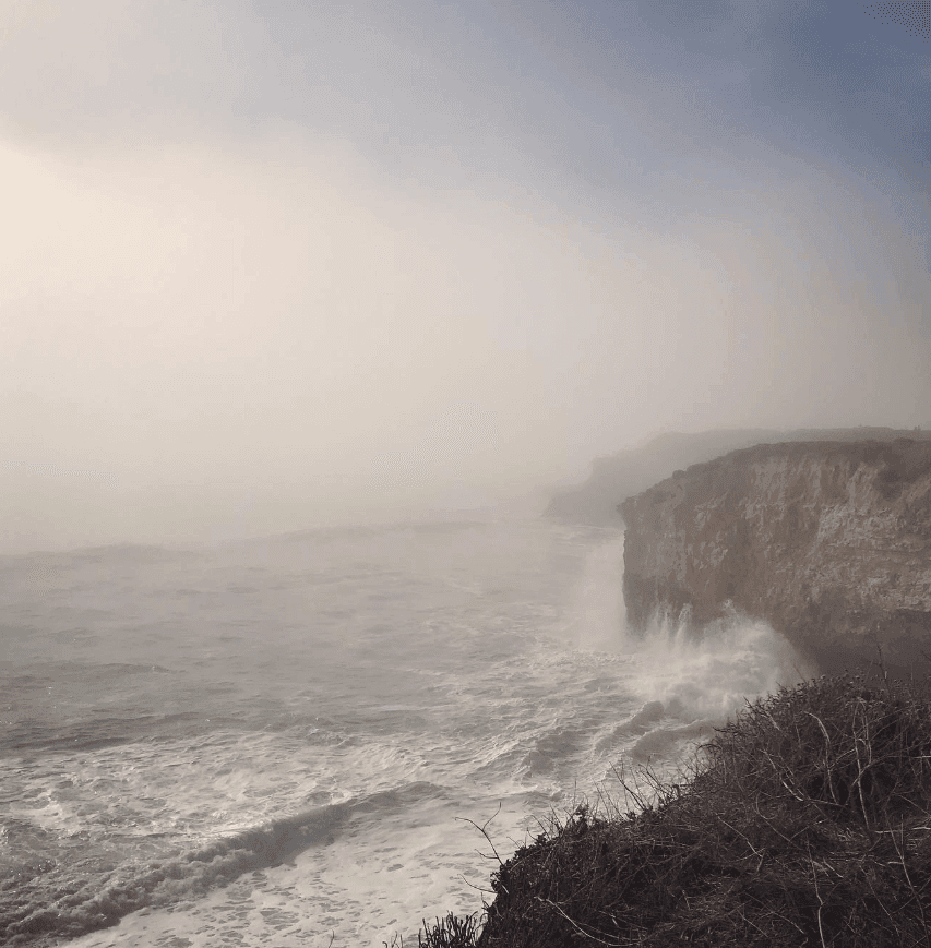 White surf breaks against steep coastal cliffs under a misty sky.