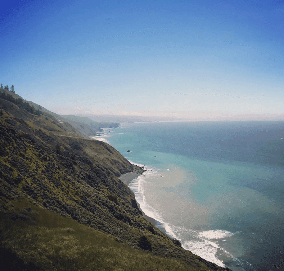 A steep green hillside drops toward turquoise water and a curving shoreline.