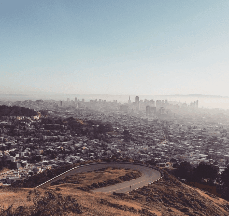 A winding hilltop road overlooks a dense city and a hazy distant skyline.