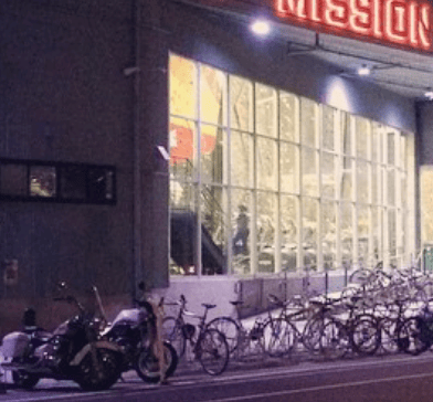 A row of bicycles lines the sidewalk outside a brightly lit storefront after dark.