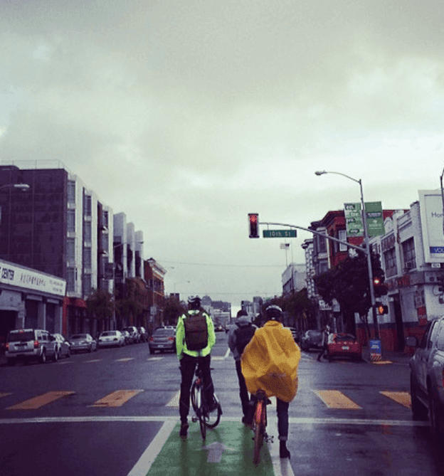 Two cyclists wait at a city intersection beneath an overcast sky.