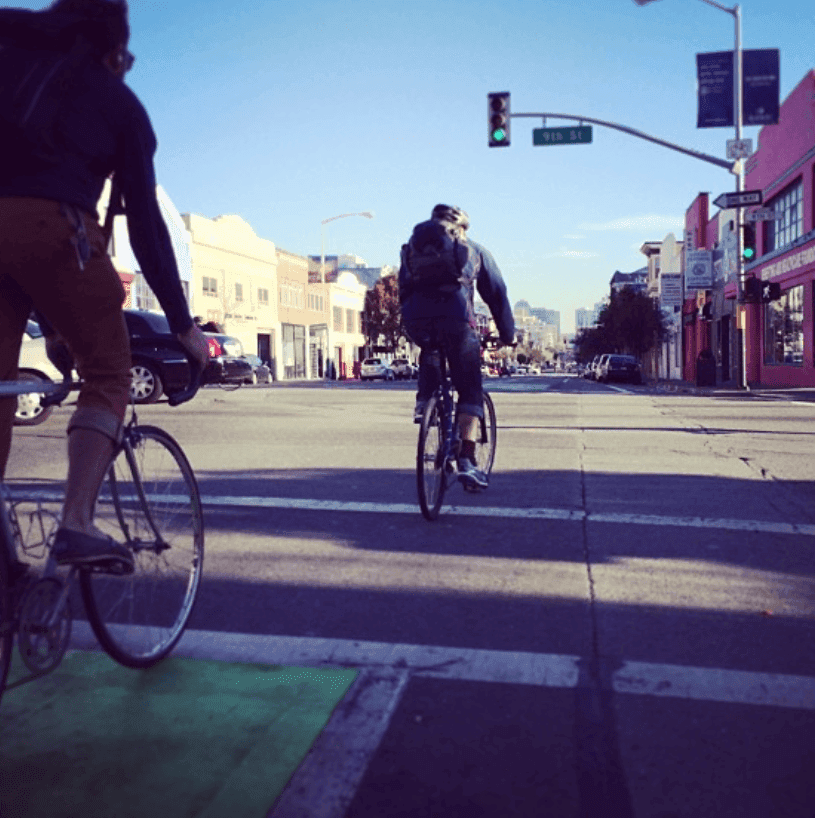 Cyclists cross a sunlit city intersection beside a painted bike lane.