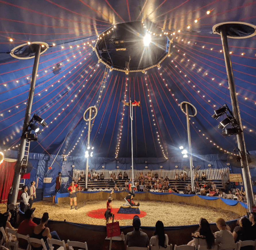 Performers and spectators gather inside a brightly lit circus tent.