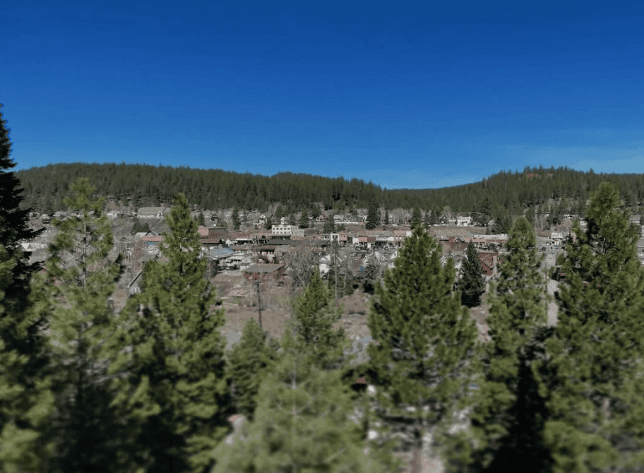 Buildings sit among evergreen trees beneath low hills and a clear blue sky.