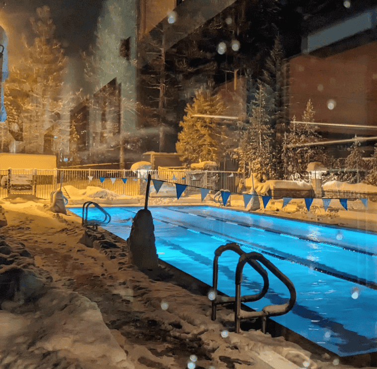 Steam rises from an illuminated outdoor swimming pool surrounded by snow.