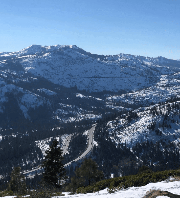 Snow-covered mountain ridges and forested valleys extend beneath a clear blue sky.